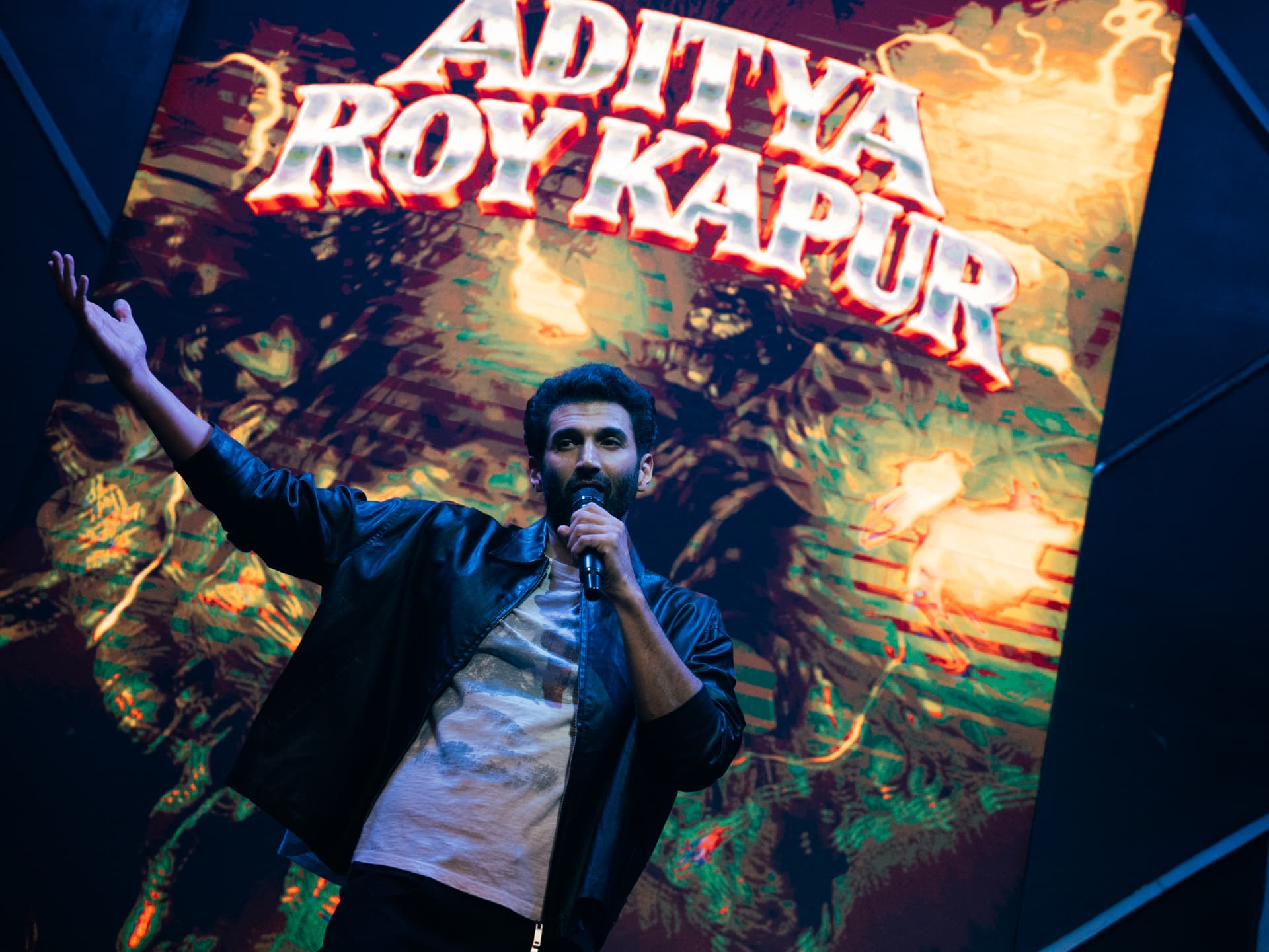 Aditya Roy Kapur raising his arm while singing beneath a blazing stage backdrop